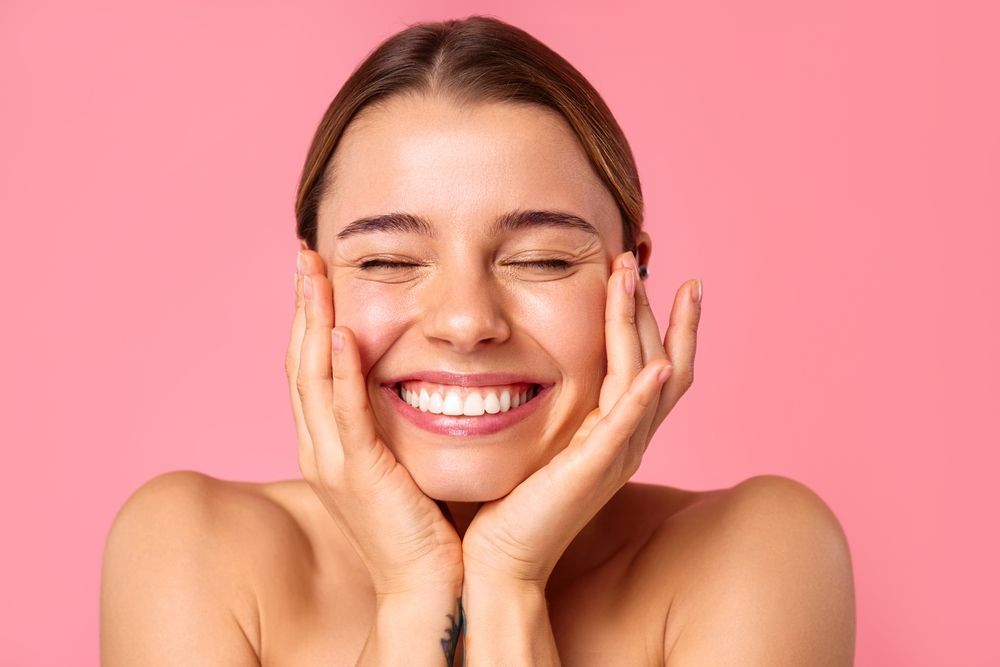 A woman is smiling with her eyes closed and her hands on her face.