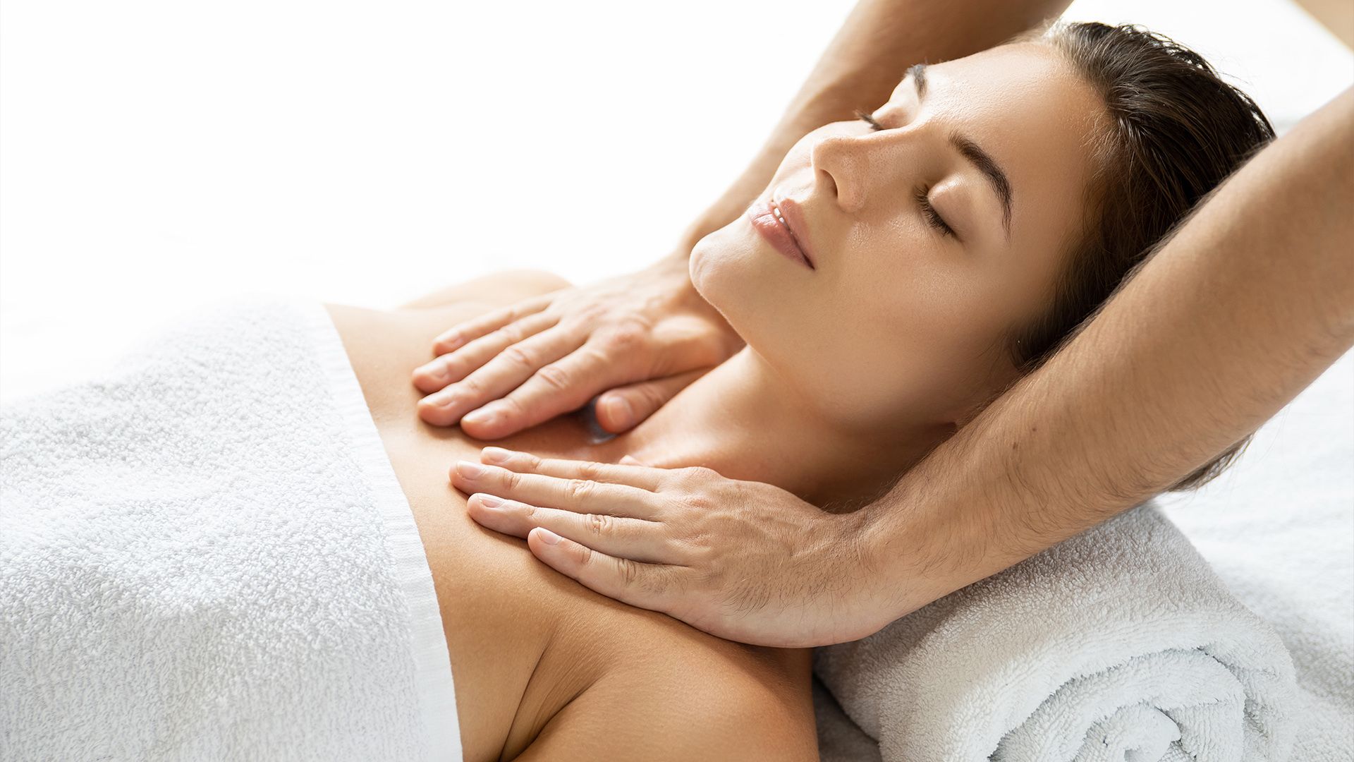 Gözleri kapalı bir kişi, masaj masasında yatarken omuzlarına ve göğsüne nazik bir masaj uygulanıyor.