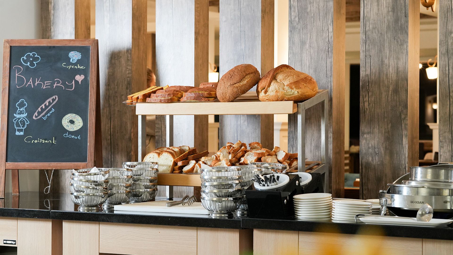Ahşap bir tezgah üzerinde taze ekmek ve pastaların sergilendiği bir fırın vitrini, yanında kara tahta tabela