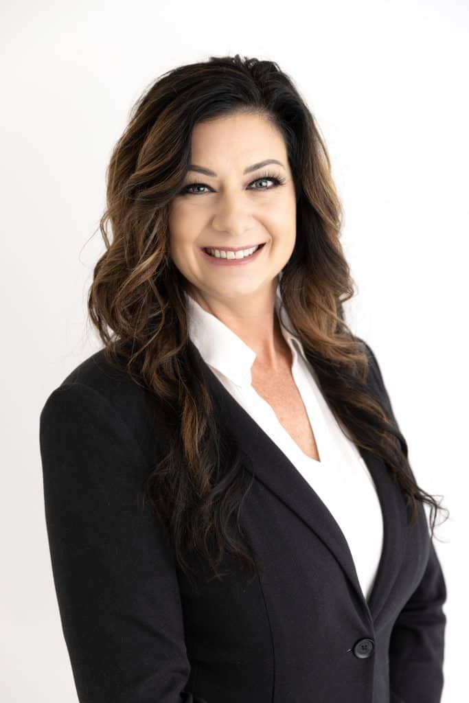 Woman smiling, wearing black suit jacket over white shirt, against a white background.