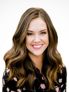 Woman with wavy brown hair smiles at the camera, wearing a dark floral shirt.