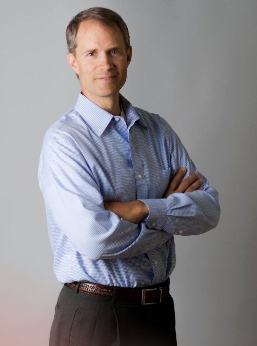 Man with arms crossed, wearing a blue shirt and brown pants, smiles in front of a gray backdrop.