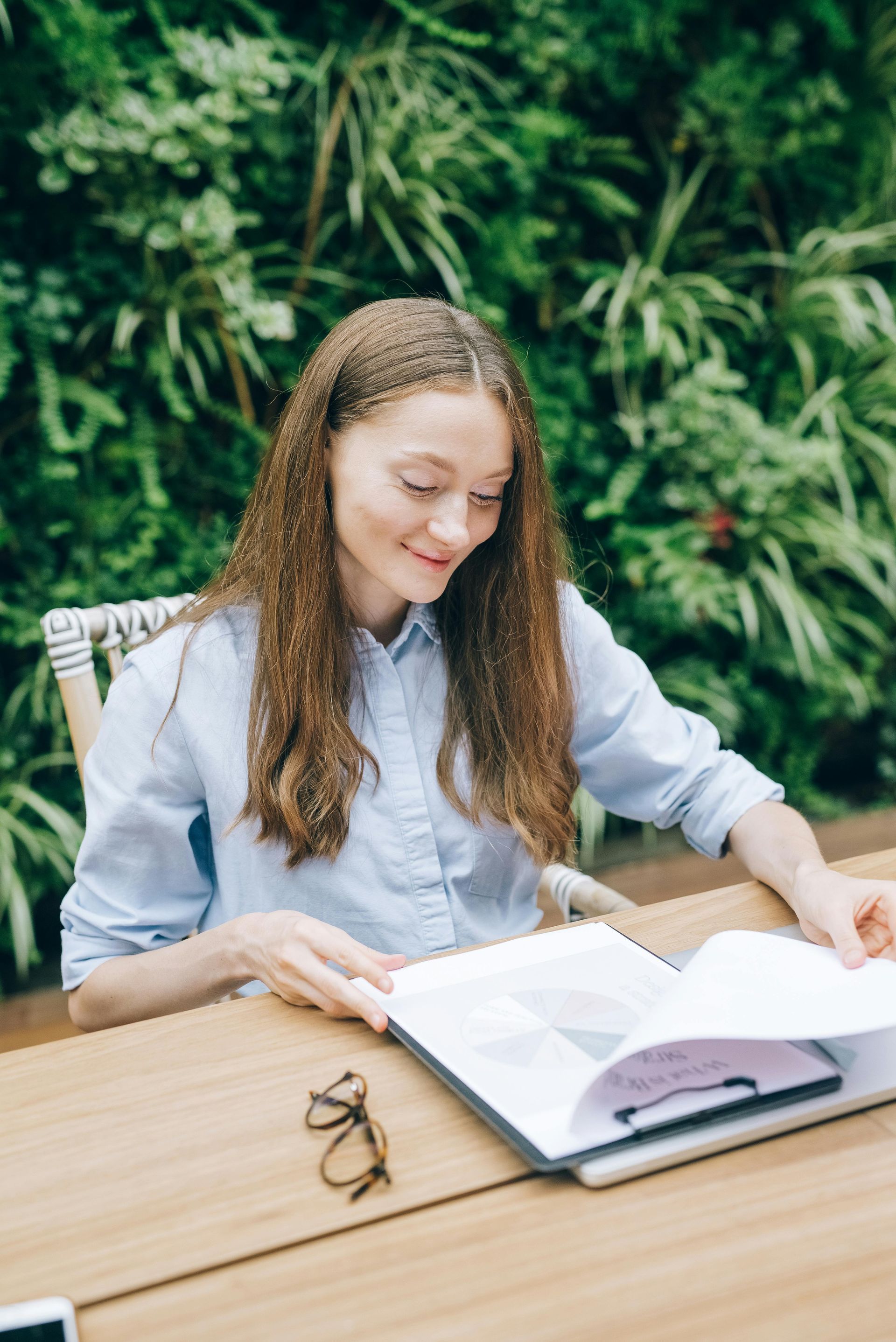 Woman with long hair reads a book at an outdoor table with greenery background. She smiles, glasses beside it.