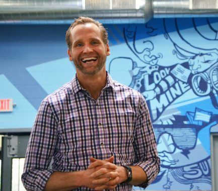 A man wearing a plaid shirt smiling in front of a wall that says look mine