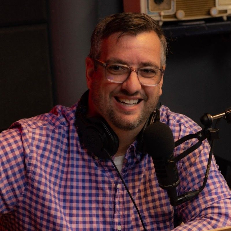 A man wearing glasses and headphones is smiling in front of a microphone.
