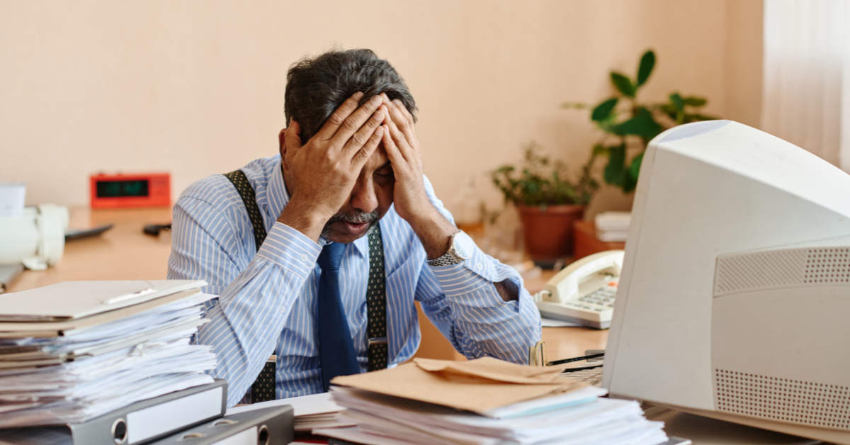 A stressed professional sitting at a cluttered desk with his head in his hands, overwhelmed by work 