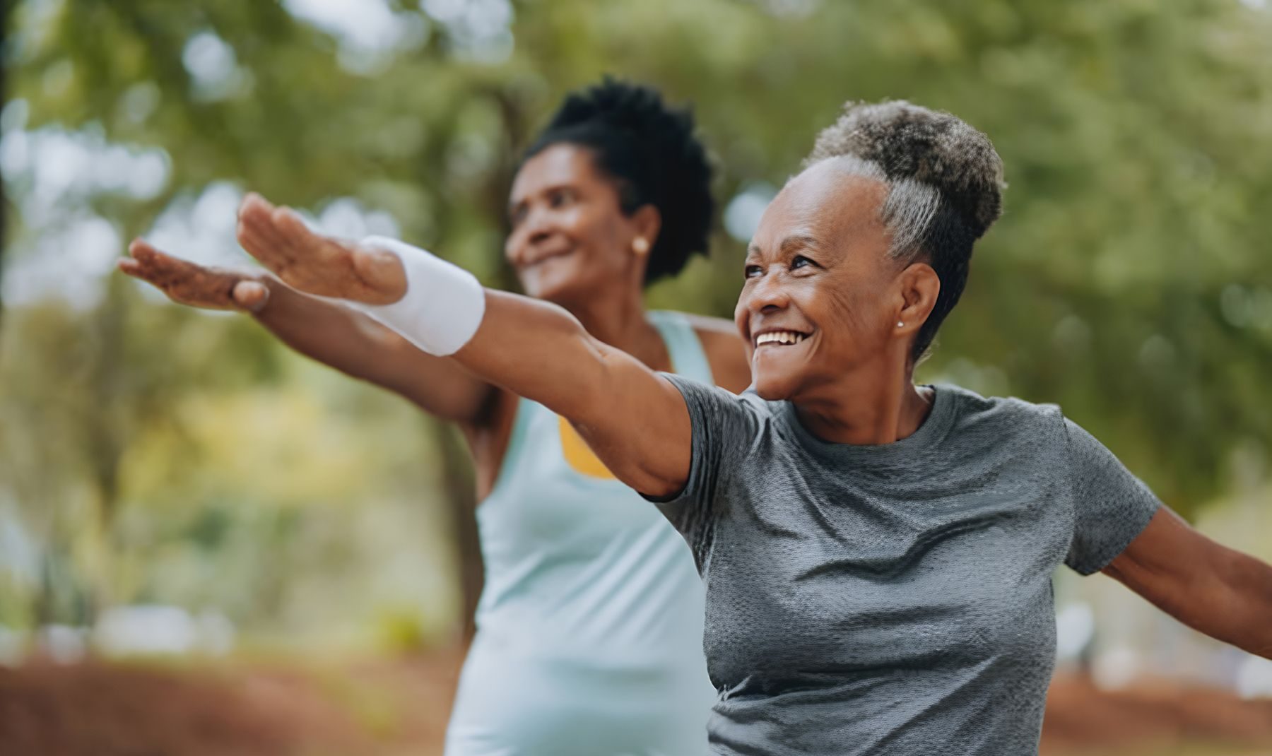 Two smiling older women exercising outdoors, arms outstretched in a park.