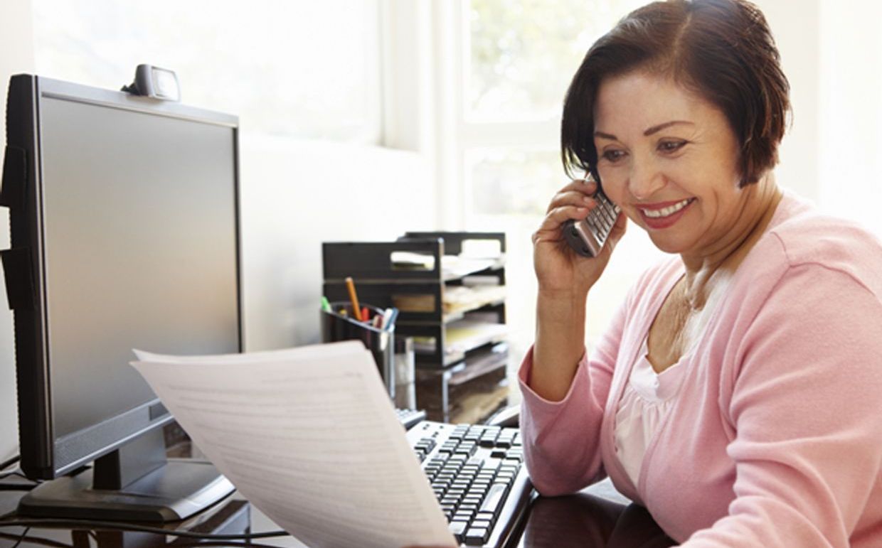 A woman is sitting at a desk talking on a cell phone while looking at a piece of paper.
