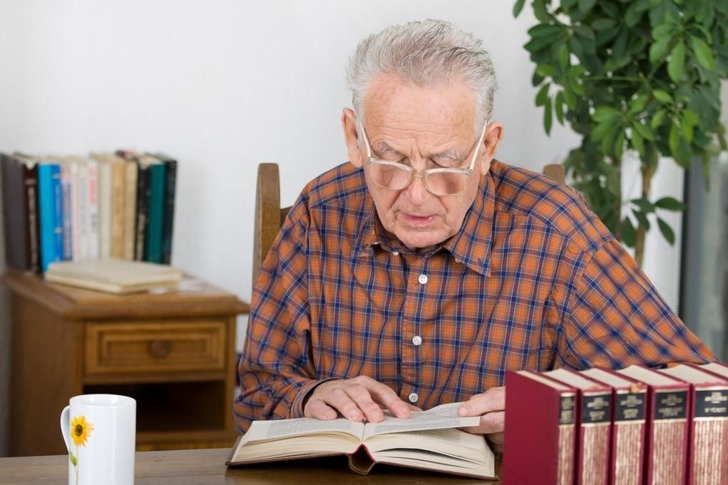 An elderly man is sitting at a table reading a book.
