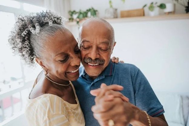 An elderly couple is dancing together in a living room.