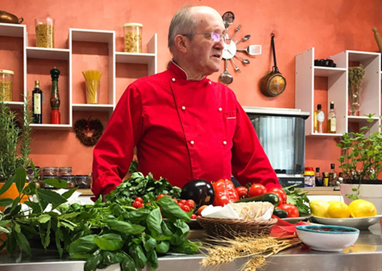 A man in a red jacket is standing in a kitchen surrounded by vegetables.
