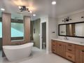 A bright, modern bathroom featuring a white soaking tub, light wood cabinetry, a double vanity, and a separate toilet room.