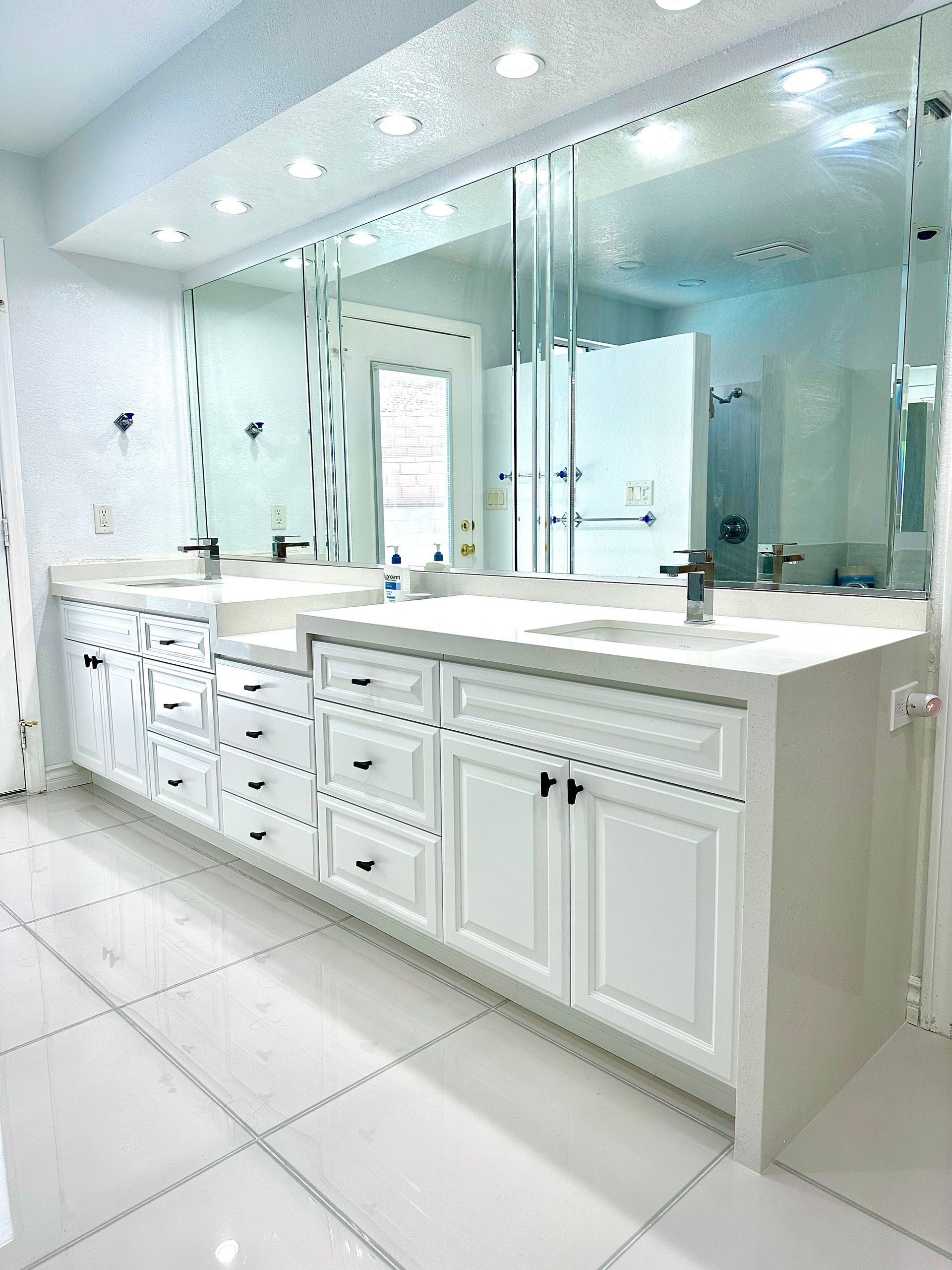 A white double vanity with silver hardware, white marble-style counters, and large mirrors in a tiled bathroom.