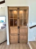 A light-wood, floor-to-ceiling built-in cabinet with glass-paned upper doors, lower panels, and vertical metal hardware.