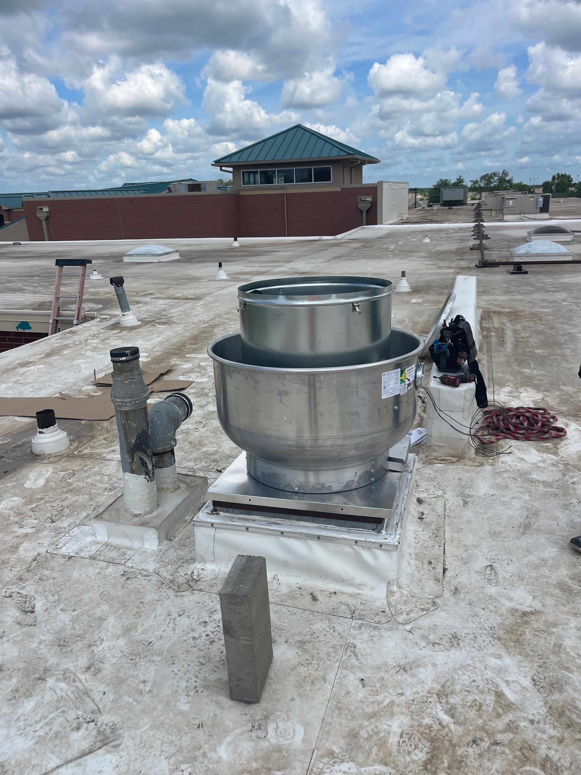 Rooftop with a large metal vent, possibly for HVAC. A building and cloudy sky are in the background.