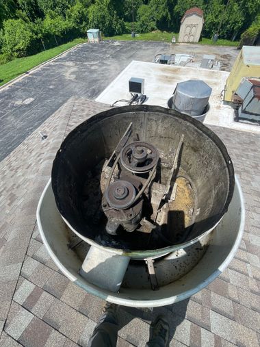 A dirty, open roof-mounted exhaust fan with a complex interior on a rooftop with foliage in the background.