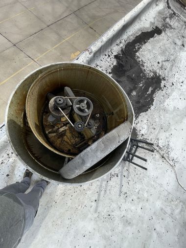 Overhead view of a cylindrical industrial fan unit on a roof, covered in black debris and an impeller blade.