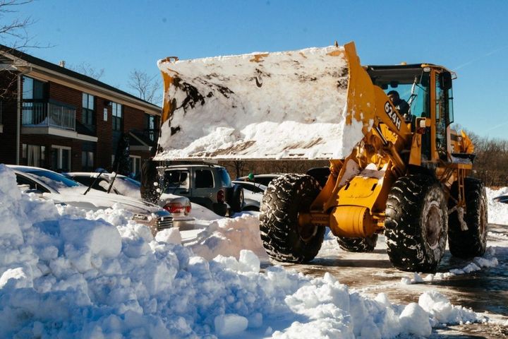 Yellow front loader clearing snow in a parking lot beside buildings, with piles of snow around it