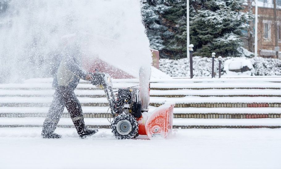 Person pushing a snow blower through heavy snowfall on a snow-covered sidewalk