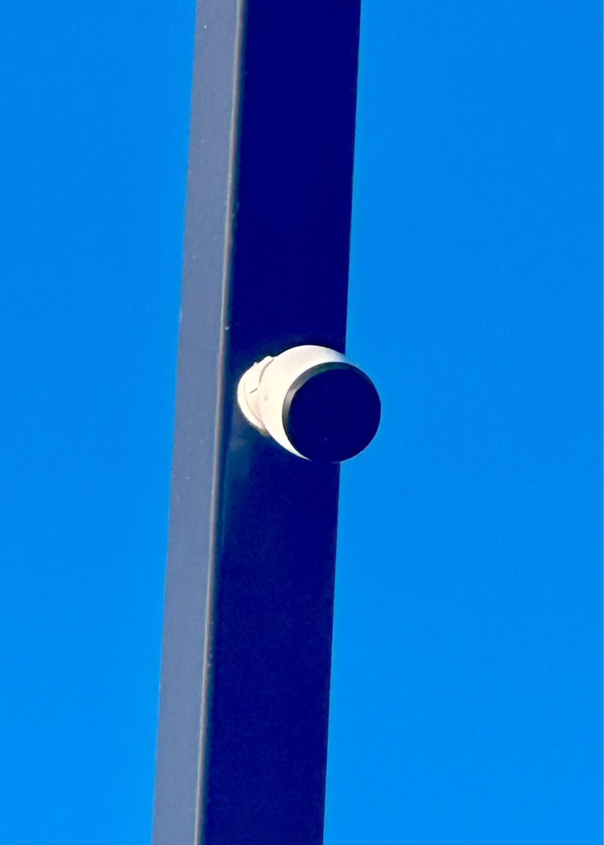 Blue metal pole with a white and black cylindrical component attached, against a clear blue sky.