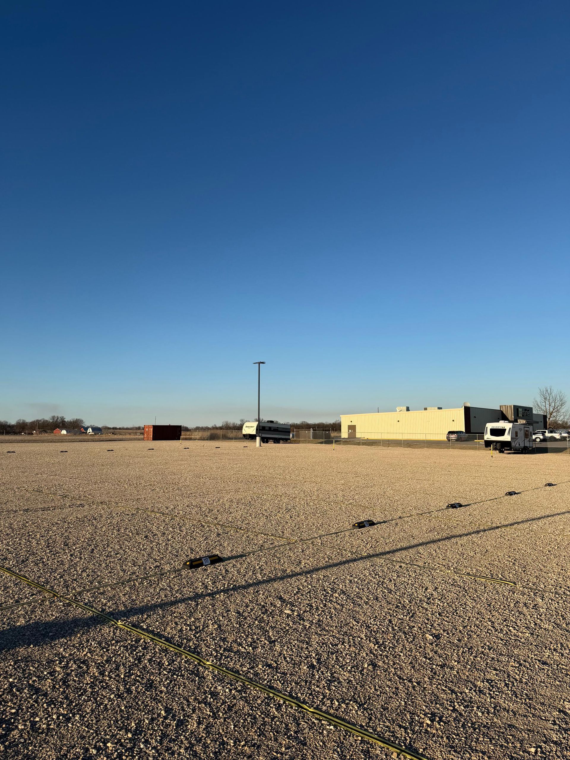 A large, gravel-covered lot under a clear blue sky, featuring a beige industrial building and distant structures.