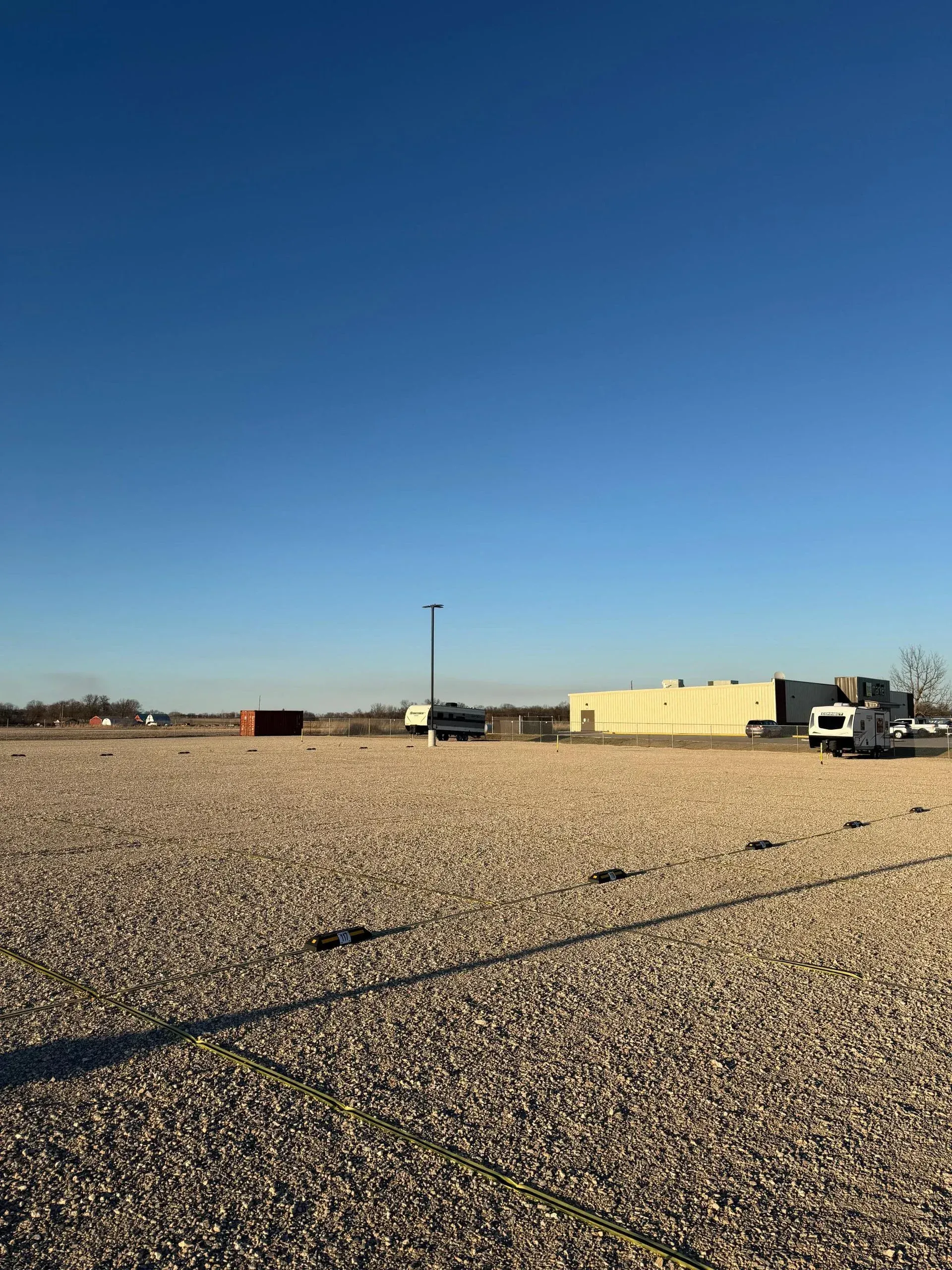 Large gravel lot with a building and vehicles under a blue sky.