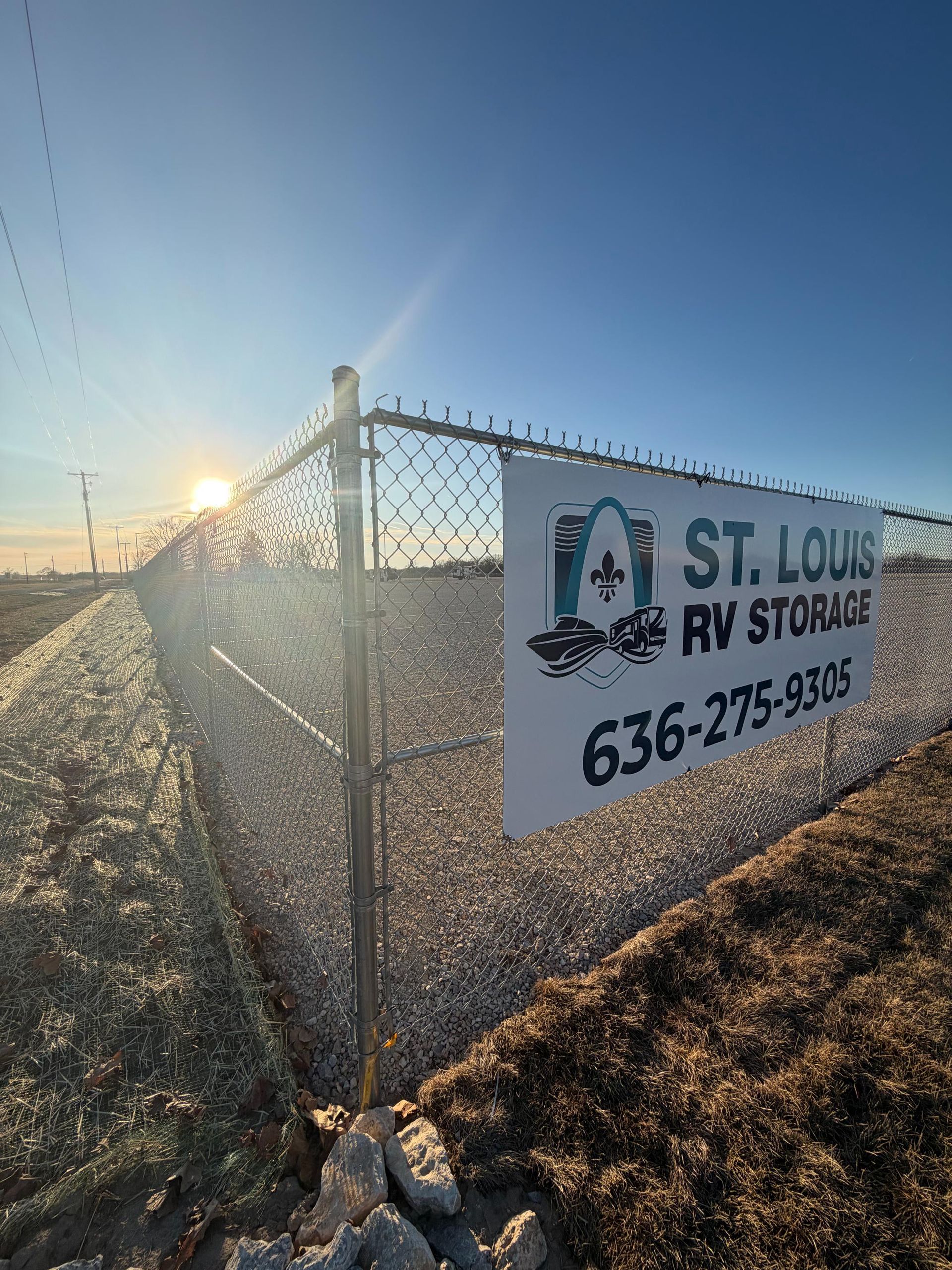 A sign for St. Louis RV Storage on a chain-link fence under a sunny sky.