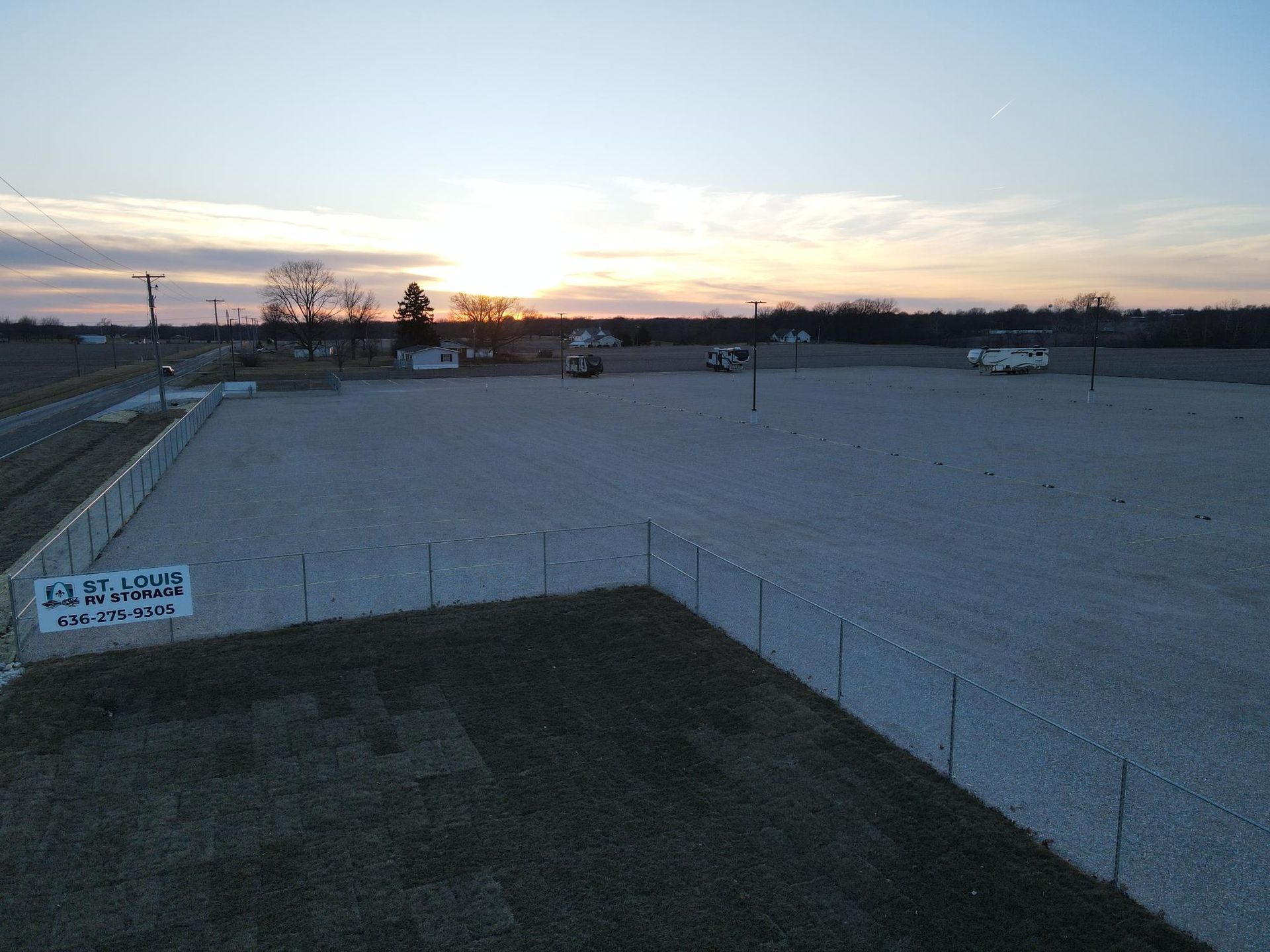An open gravel lot at sunset, with a fence, small buildings, and a few vehicles.