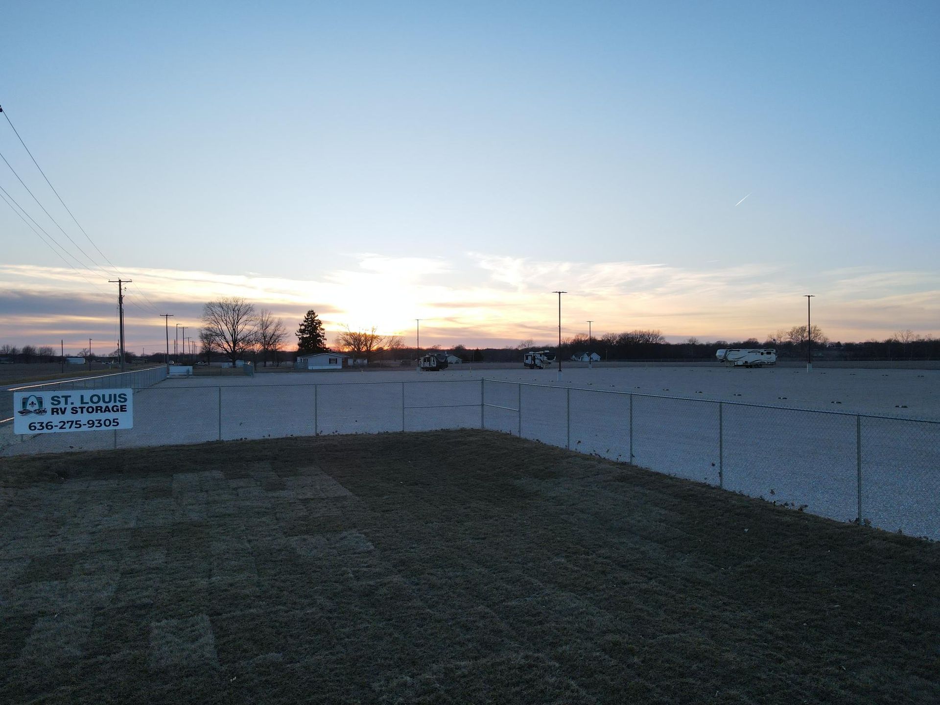 A wide, gravel parking lot at sunset with a chain-link fence and a small sign in the foreground.