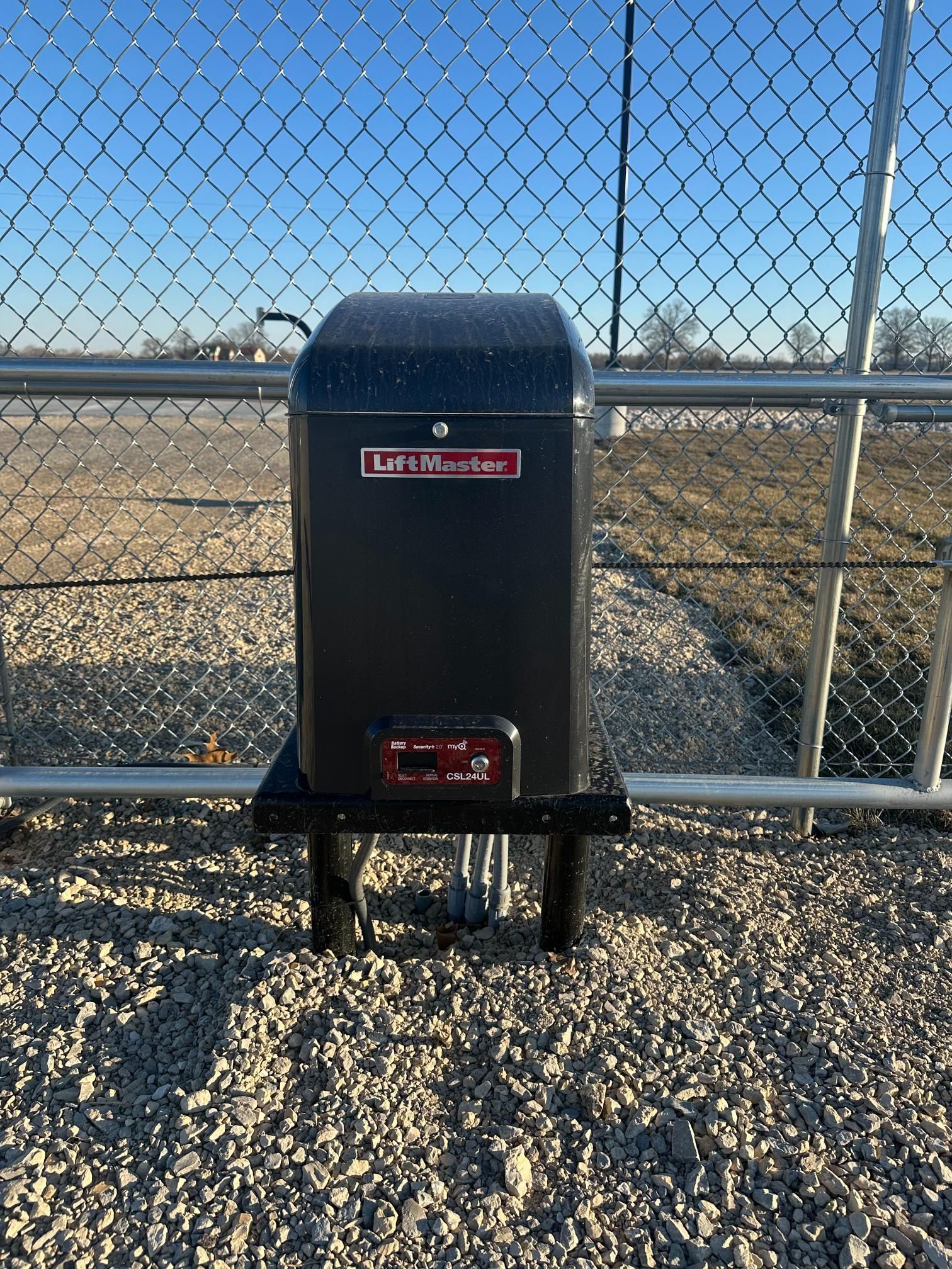 A black LiftMaster gate operator motor installed on a post in a fenced gravel lot under a clear blue sky.