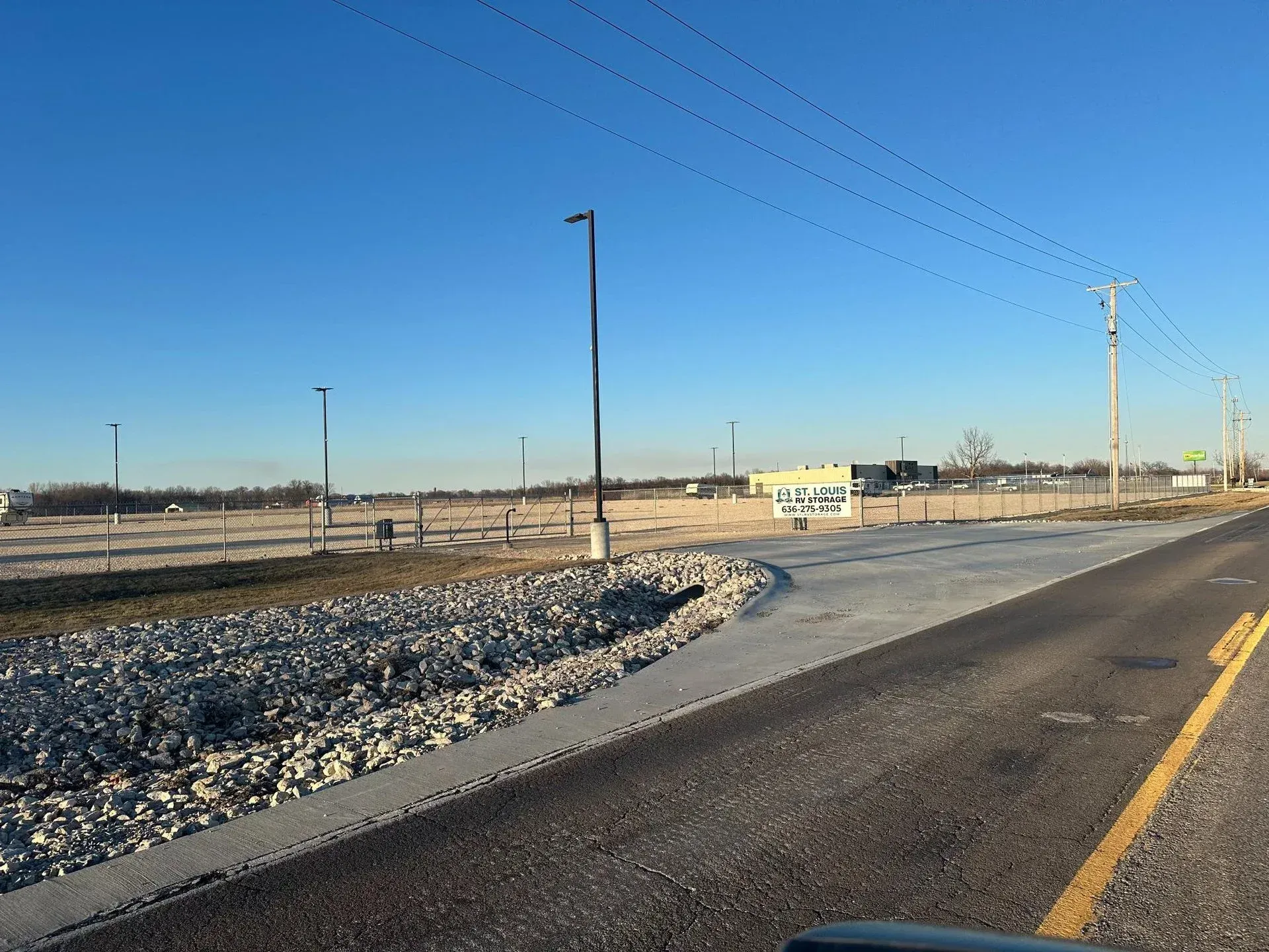 Paved road leading to a gravel area with empty lot in the distance, light poles, power lines, and clear blue sky.