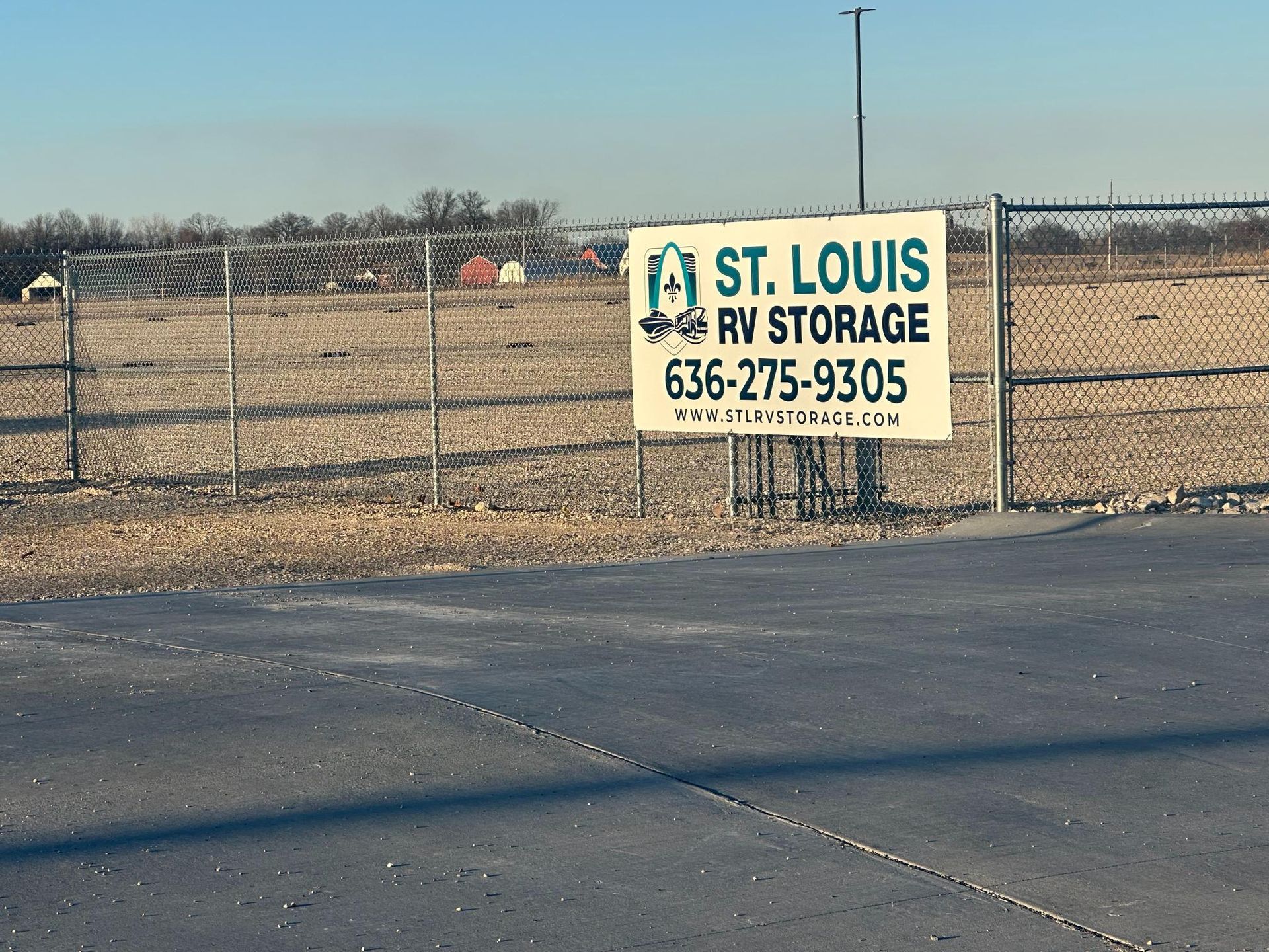 A white sign for St. Louis RV Storage with its phone number, attached to a chain-link fence bordering a gravel lot.