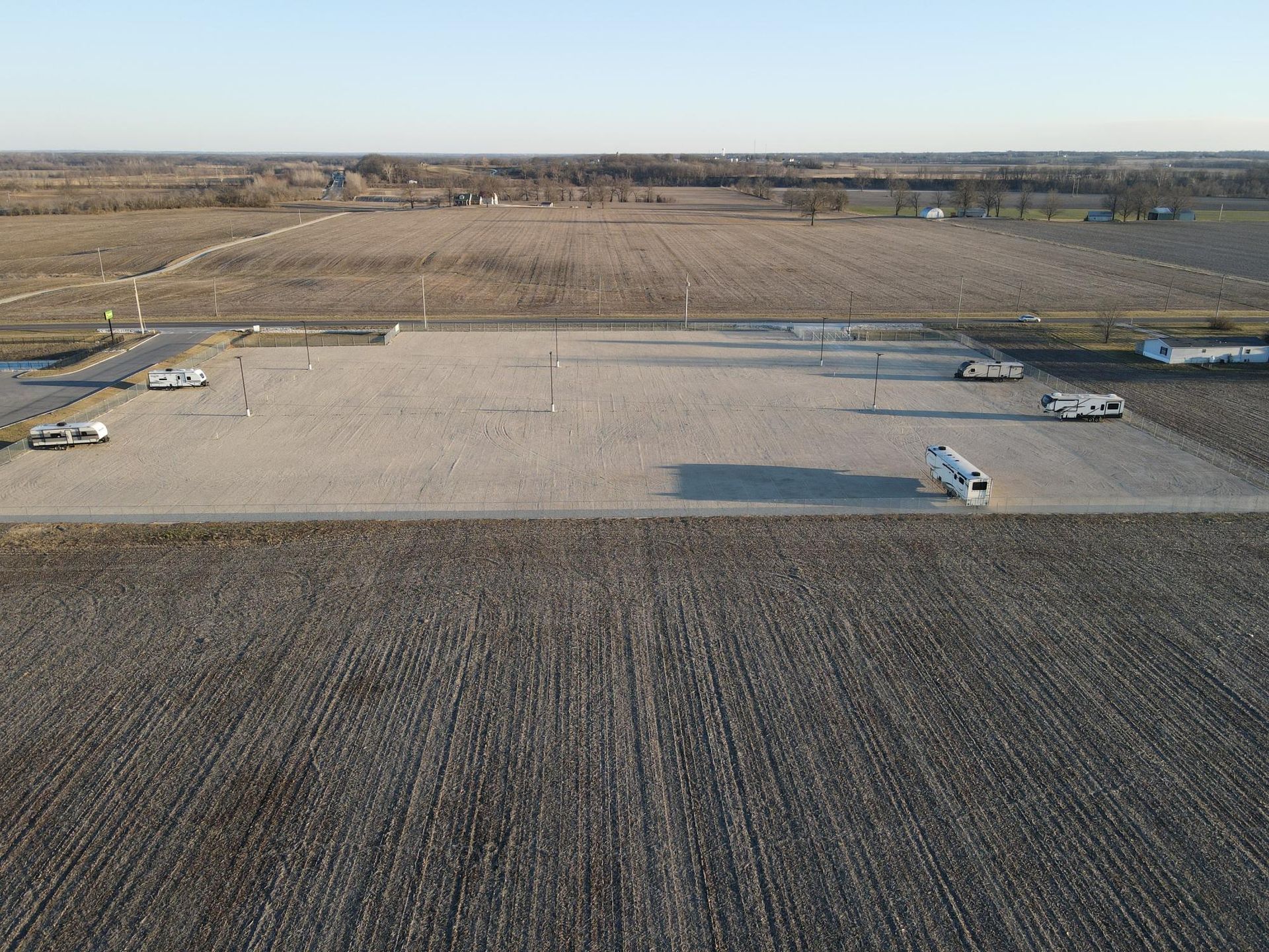 Aerial view of a gravel parking lot with a few recreational vehicles, surrounded by large, empty agricultural fields.