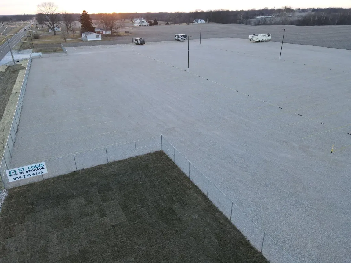 Gravel parking lot at sunset, fenced perimeter, small buildings in background, sign in lower left corner.