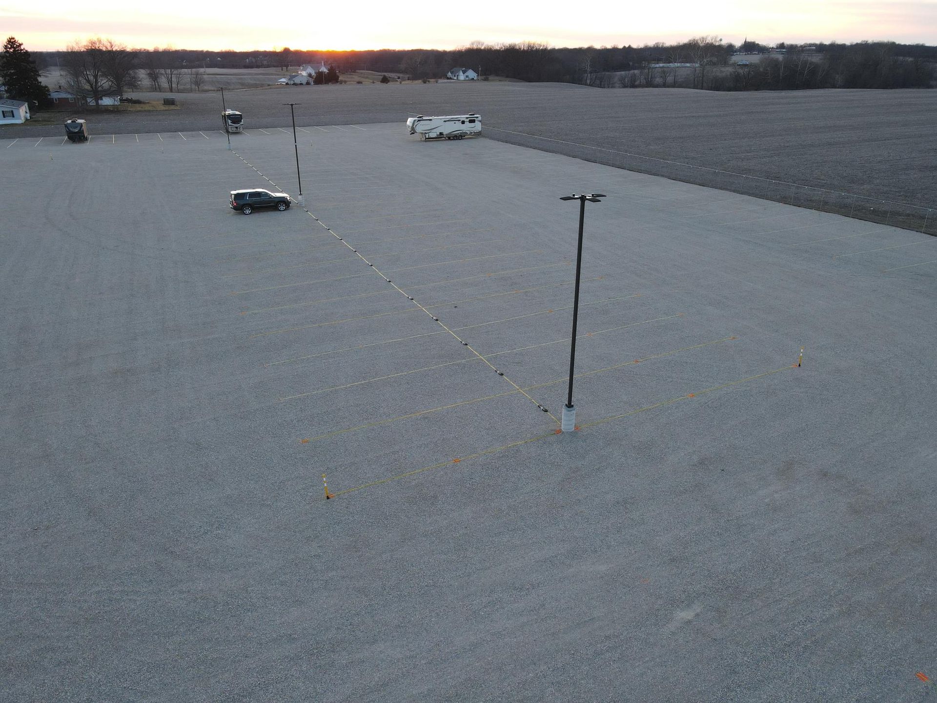 Gravel lot with a car, poles, and a camper. The sun sets in the background over a field.