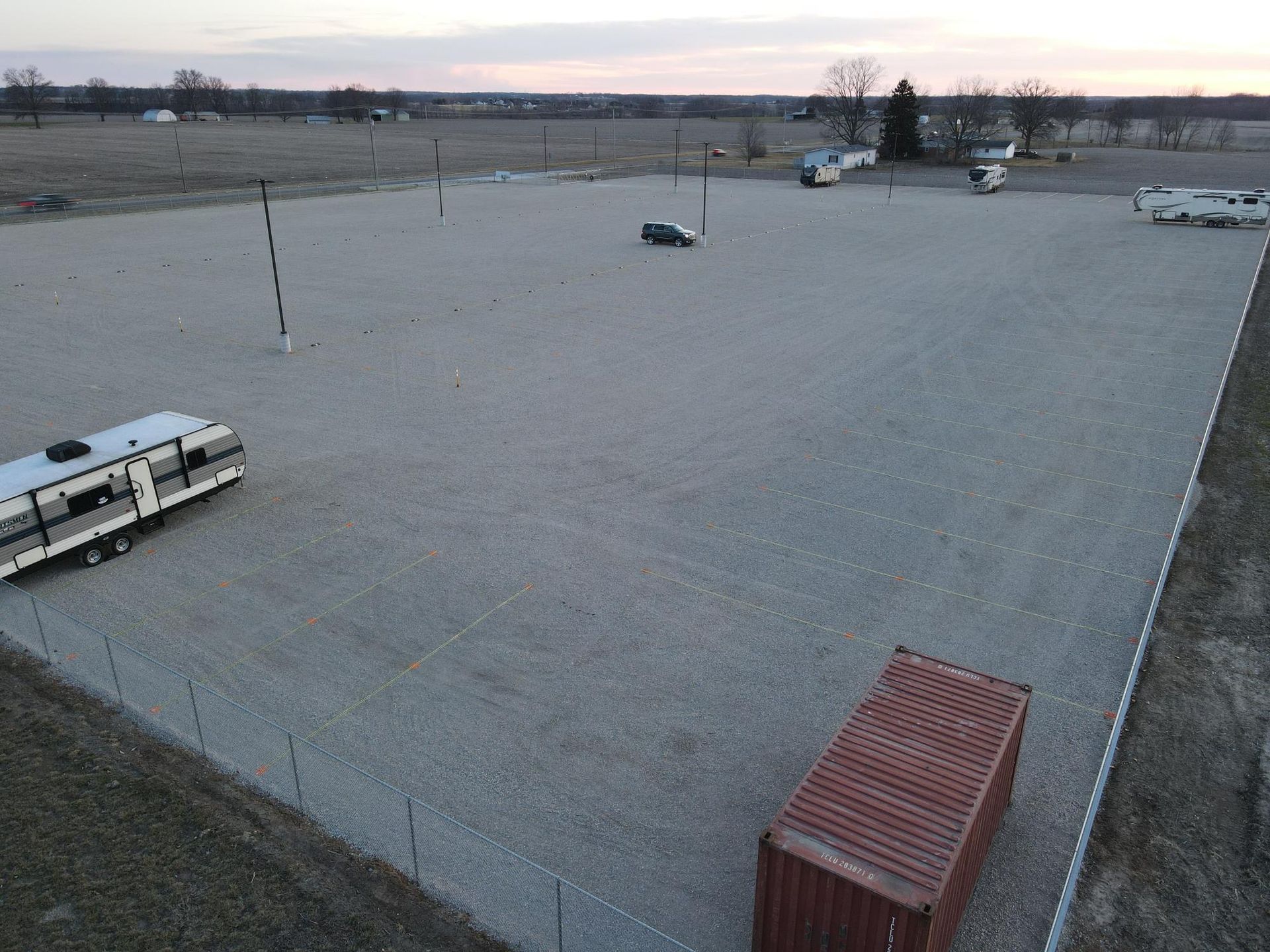 Aerial view of a gravel lot containing a travel trailer, shipping container, and a few parked vehicles.