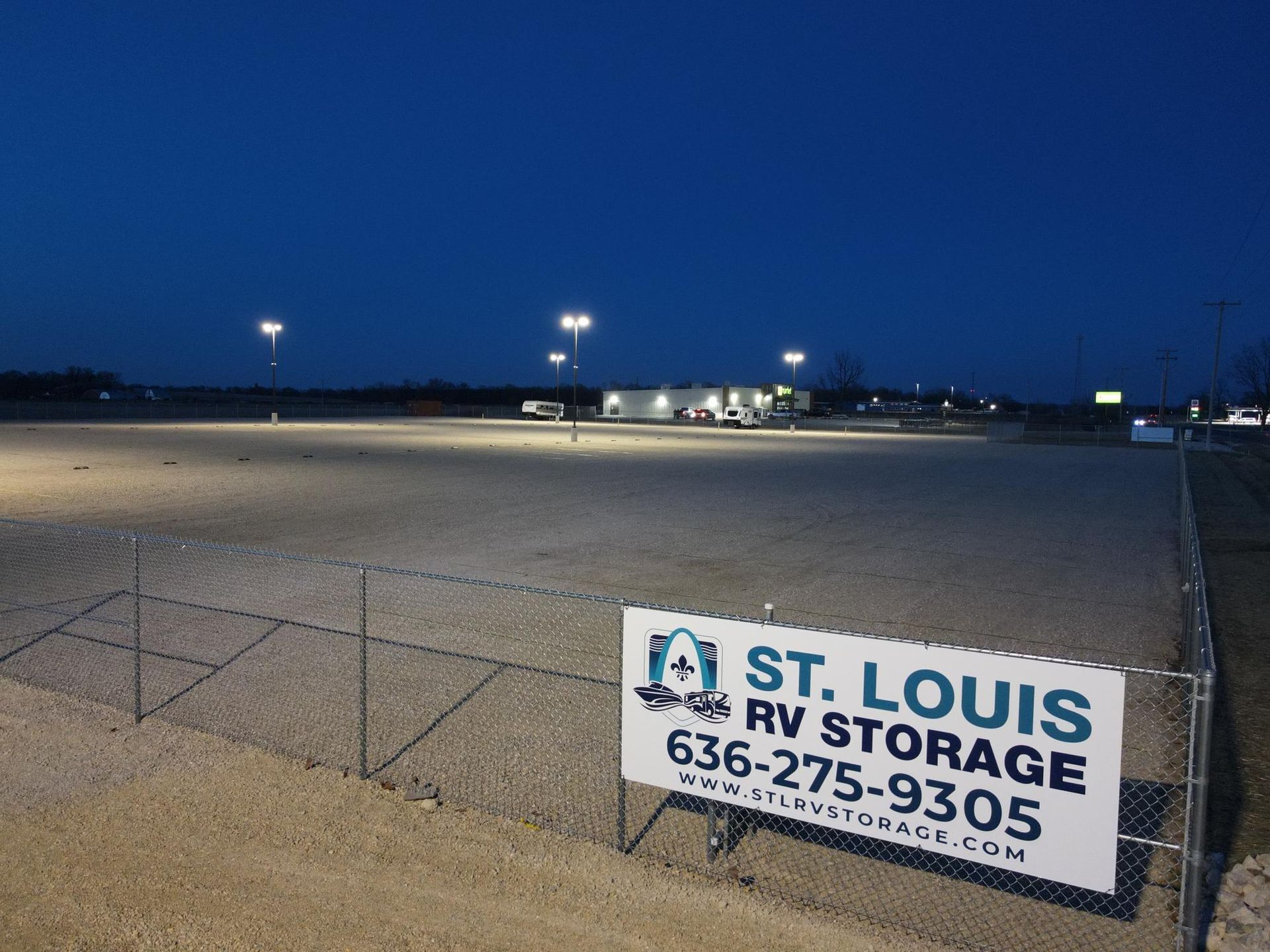 St. Louis RV Storage sign on a chain-link fence, overlooking a lit, gravel storage lot at dusk.