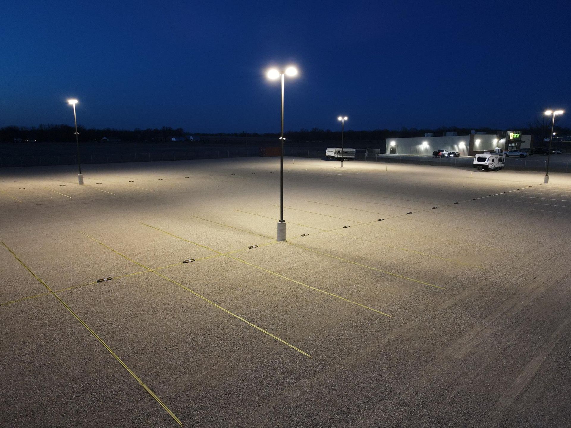 A gravel parking lot illuminated by tall LED light poles under a twilight sky.