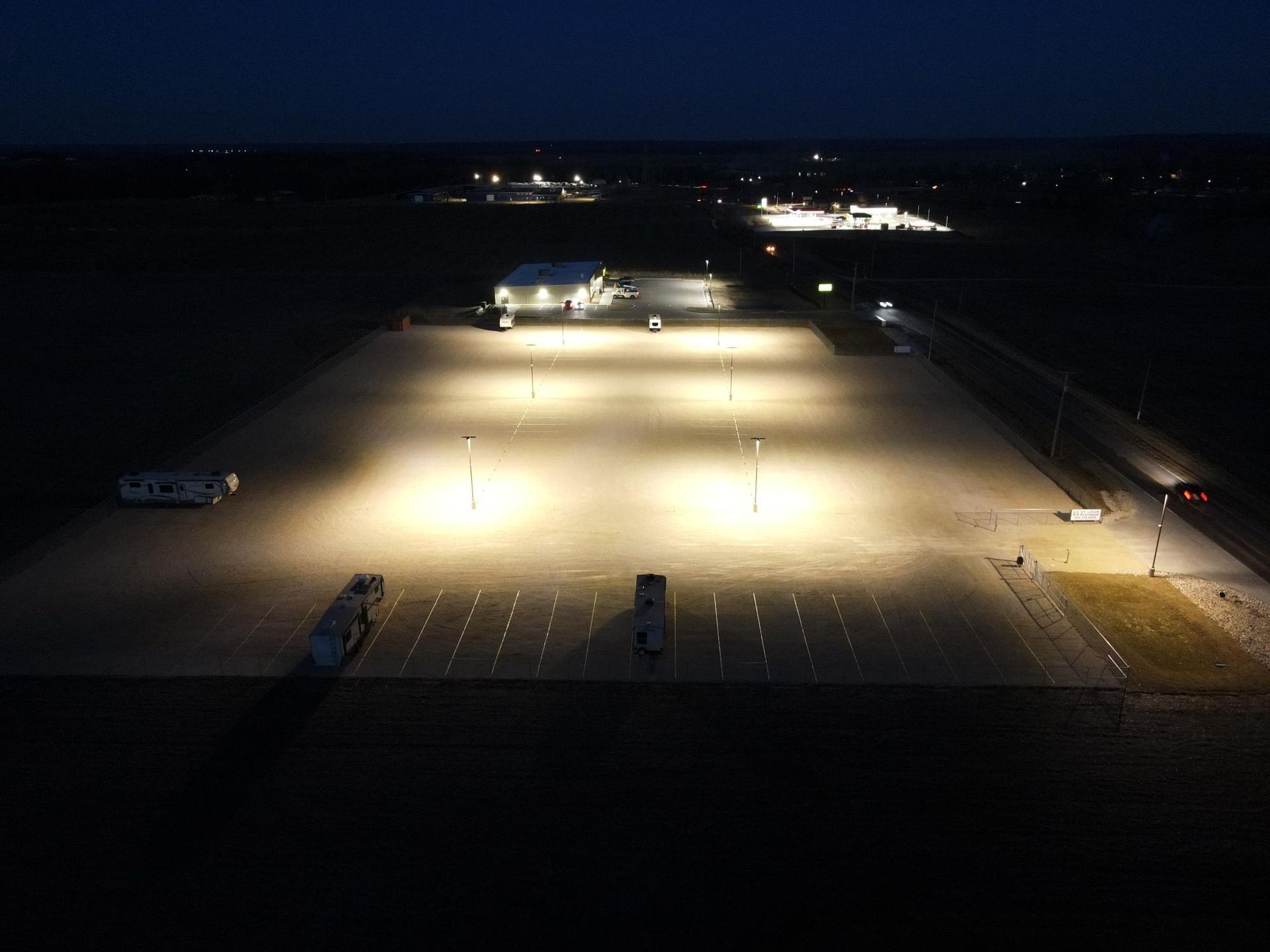 Night view of an illuminated parking lot and surrounding buildings, with a few vehicles.