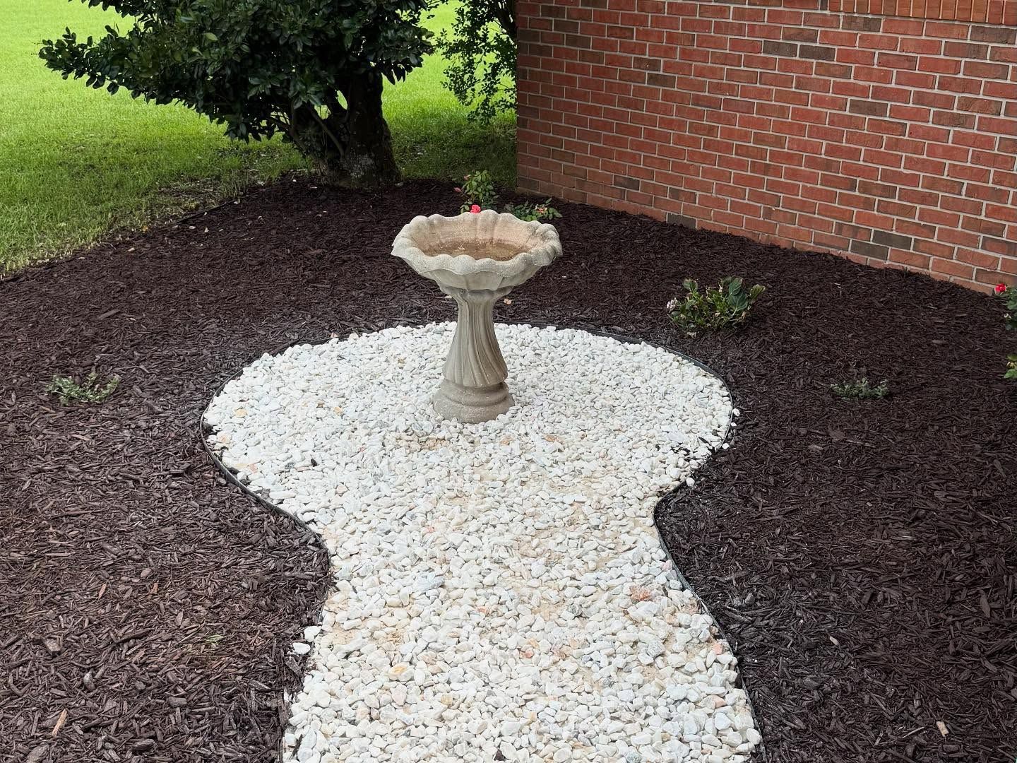 Birdbath centered in a white pebble garden path beside dark mulch, grass, and a brick wall.