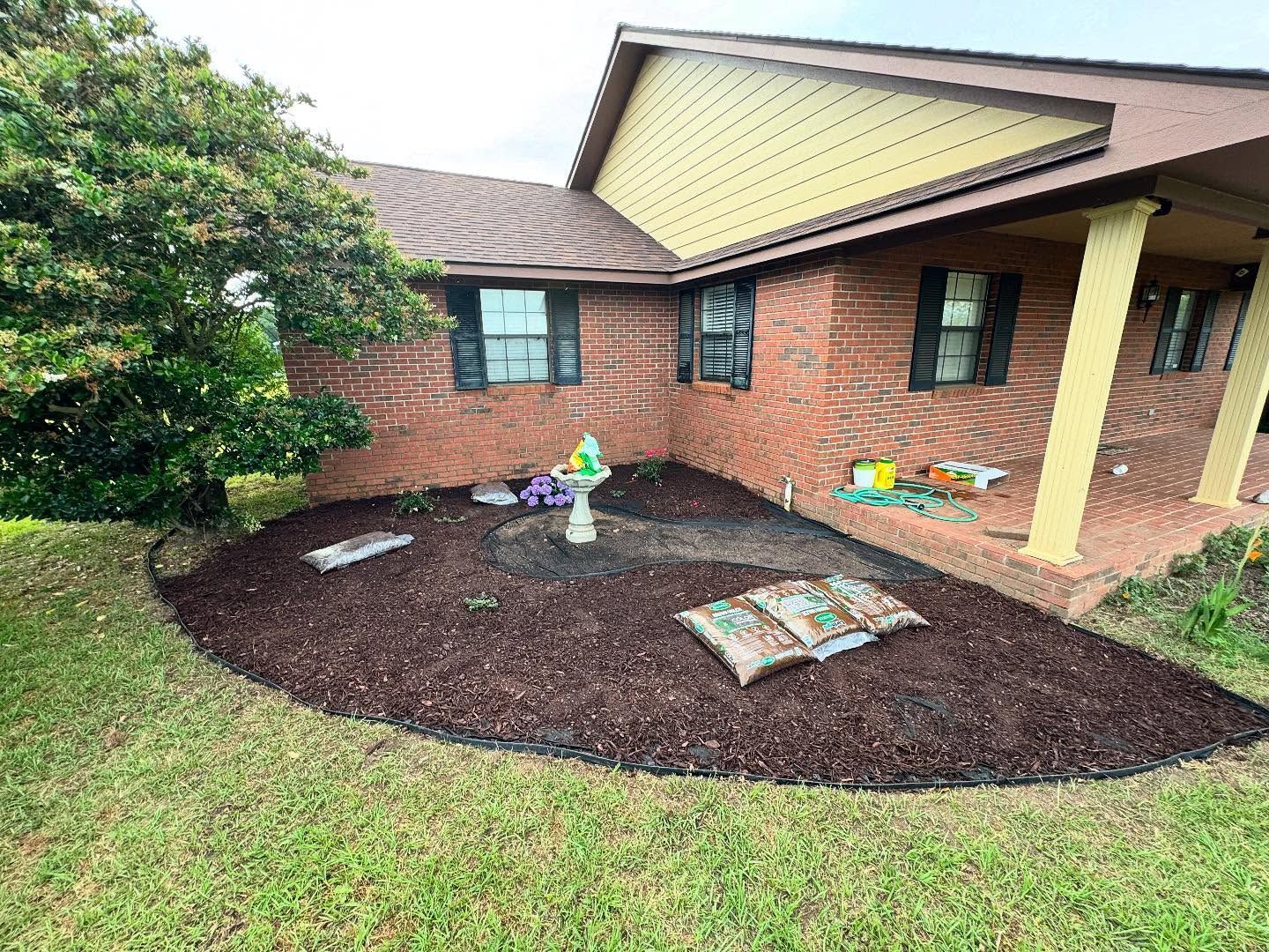 Front yard garden bed with mulch, small statues, and a brick house with a covered porch.