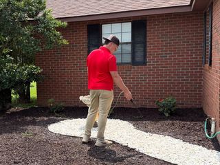 Person in a red shirt and hat walking on a gravel path beside a brick house.