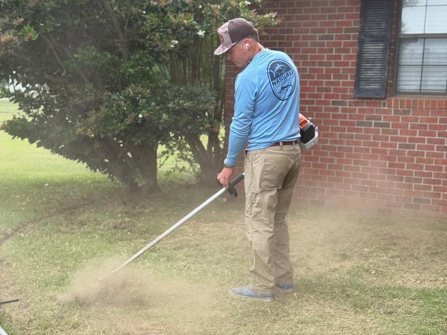 Person using a string trimmer on a lawn beside a brick house, kicking up grass clippings.