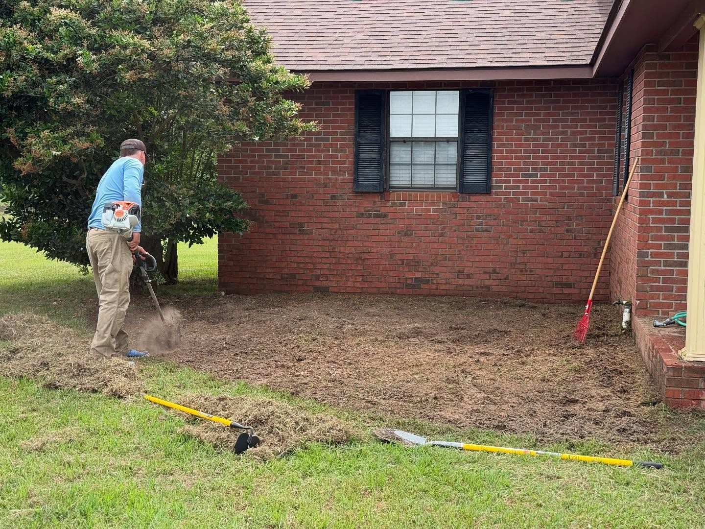 Worker raking debris beside a brick house, with a shovel and yellow hose in the yard.