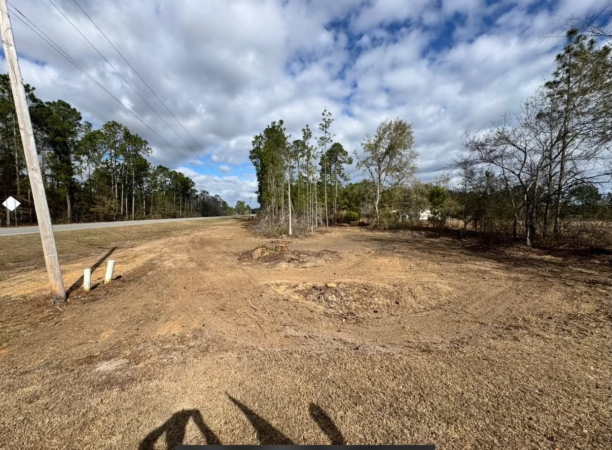 Open dirt lot beside a road under a cloudy sky, bordered by sparse trees and utility poles