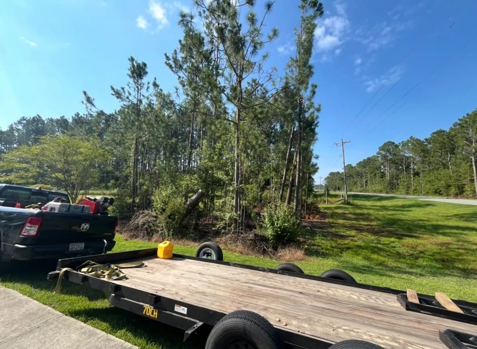 Flatbed trailer beside a black pickup on a grassy roadside, with trees and blue sky in the background.