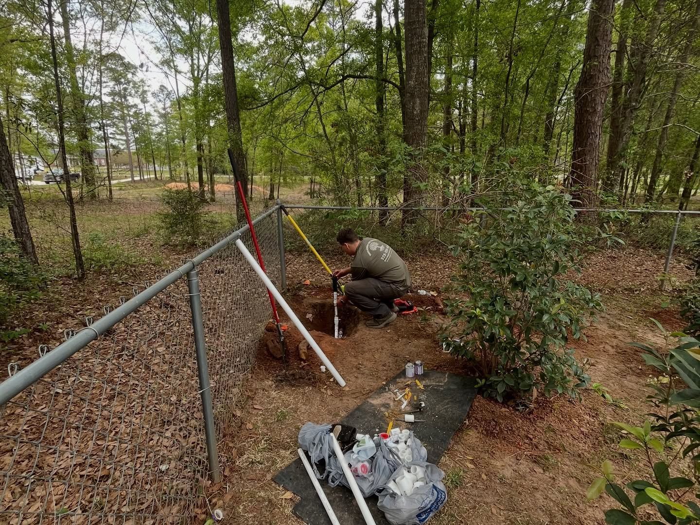 Worker in a wooded area repairing a metal fence beside a ditch with debris and water