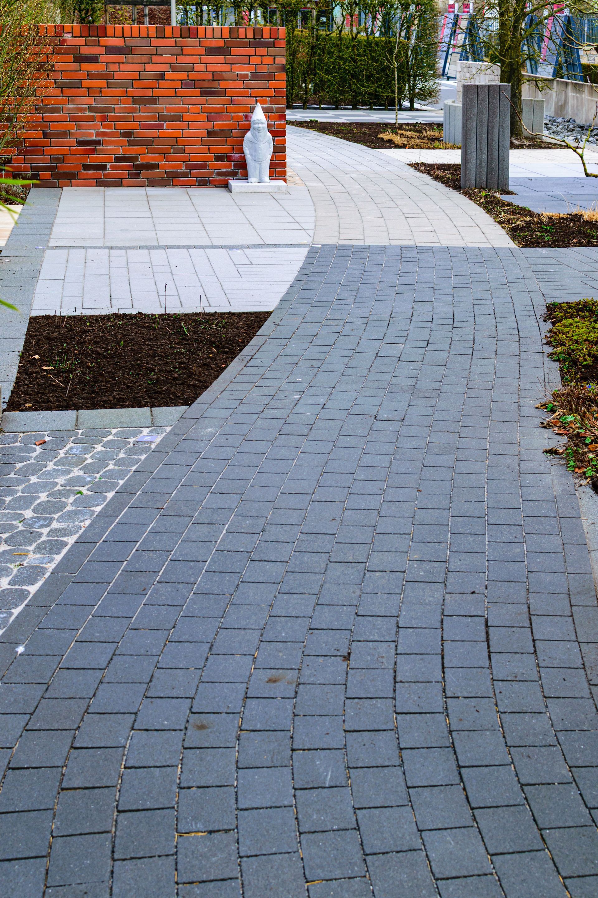 Brick-paved walkway with white stone insets beside a red brick wall and a few bollards, in a landscaped area