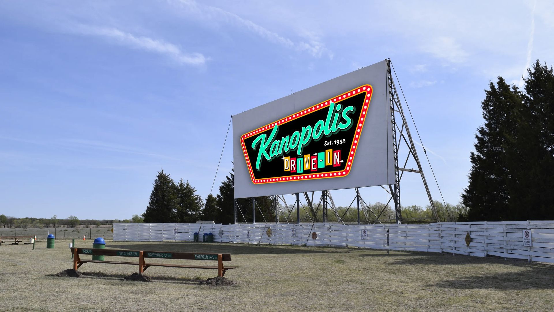 Kanopolis Drive-In movie screen with a neon sign, under a blue sky. A bench sits in the foreground.