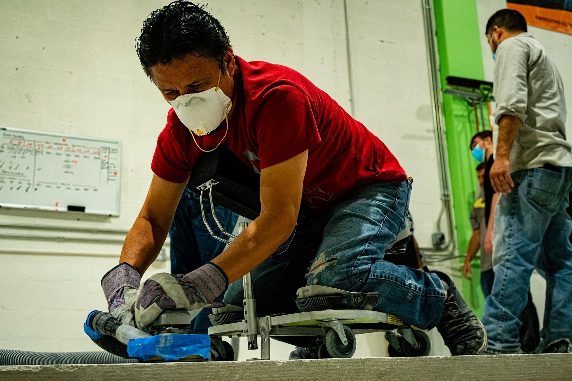 Worker in red shirt and mask kneels while smoothing concrete on a floor with a trowel.